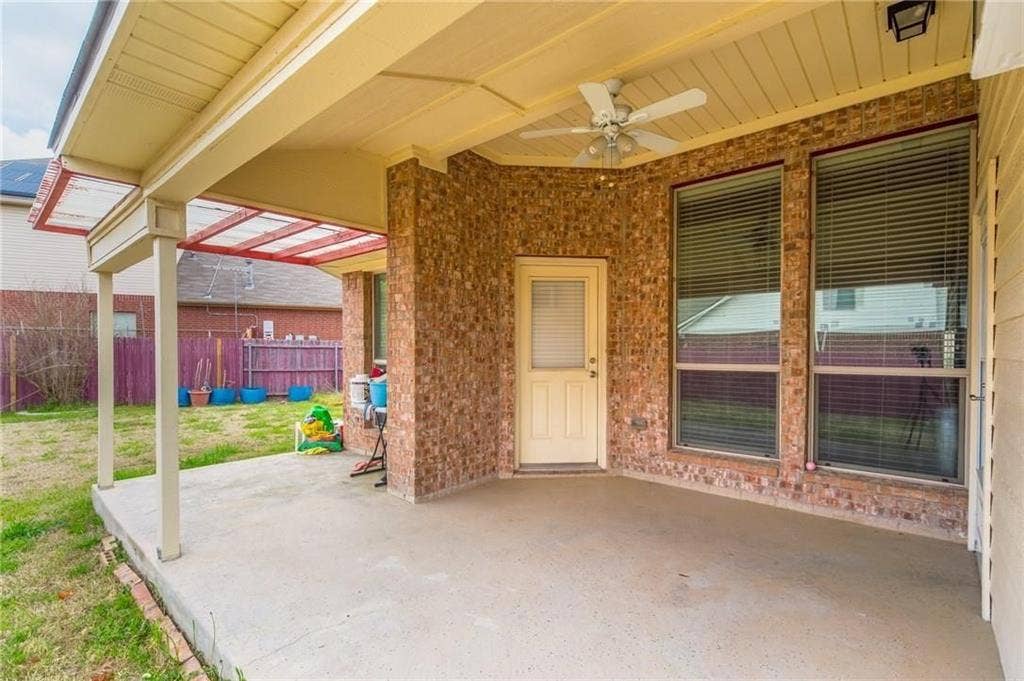 Covered outdoor patio featuring a ceiling fan, brick exterior, and two large windows with blinds