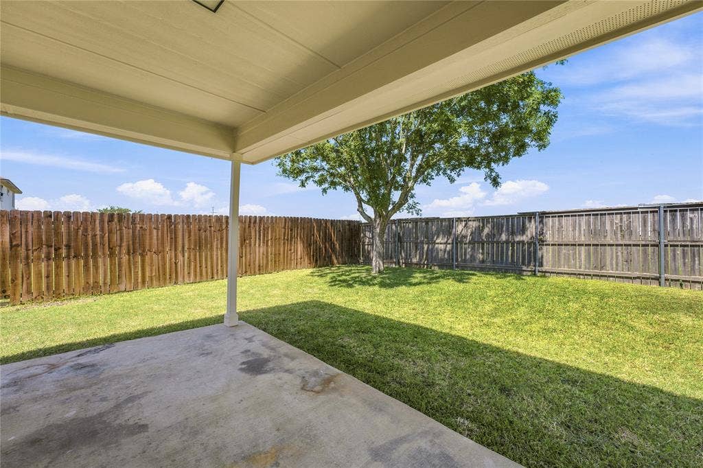 Covered concrete patio overlooking a private backyard with a mature tree, wood fencing, and manicured lawn