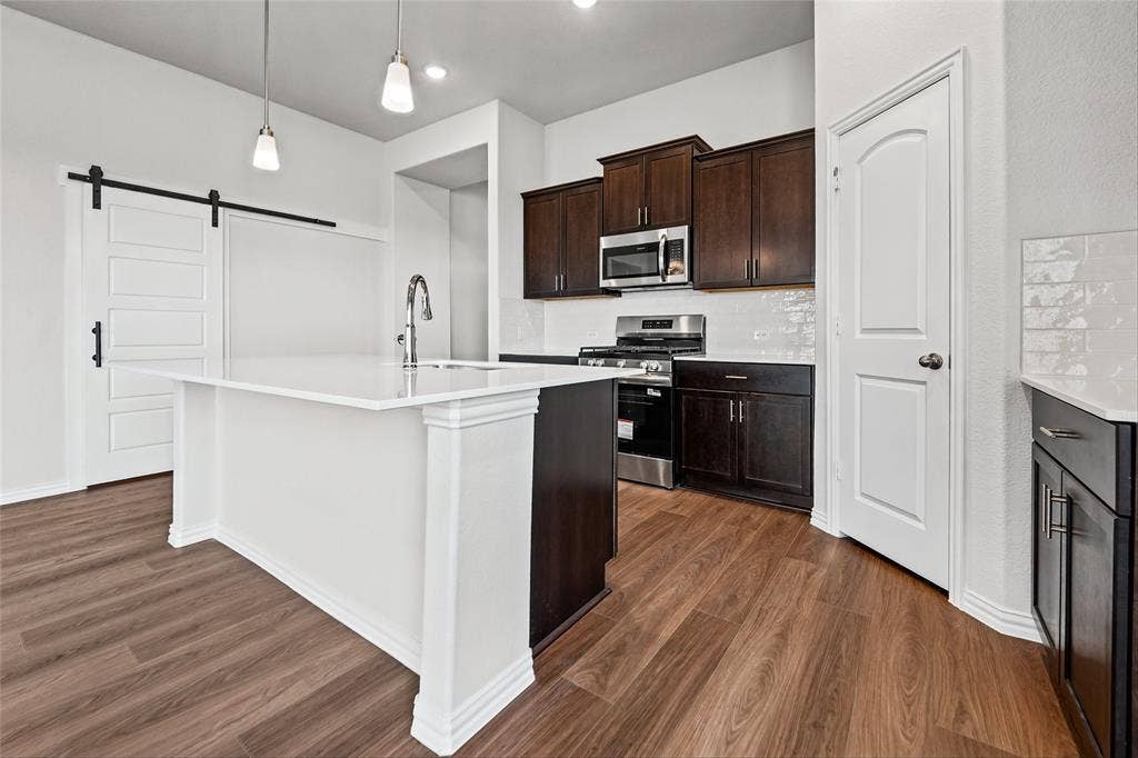 10008 Birchpoint Drive, Fort Worth, TX 76108 Kitchen featuring a center island with a sink, pendant lighting, wood-finish flooring, dark wood cabinetry, and stainless steel appliances