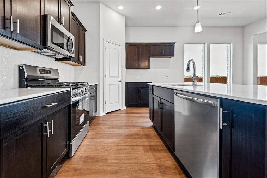 10008 Birchpoint Drive, Fort Worth, TX 76108 Modern kitchen featuring dark wood-finish cabinetry, stainless steel appliances, a center island with a sink, white countertops, and wood-finish flooring