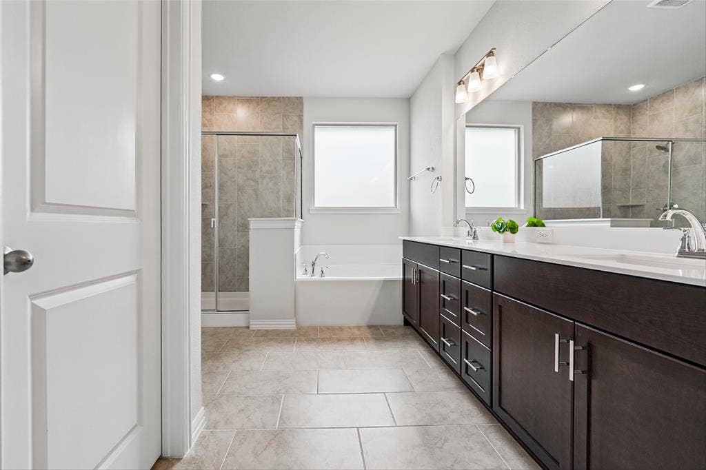10008 Birchpoint Drive, Fort Worth, TX 76108 Dual dark wood-finish vanity with white countertops, tiled flooring, built-in soaking tub, and a glass-enclosed shower with tiled surround