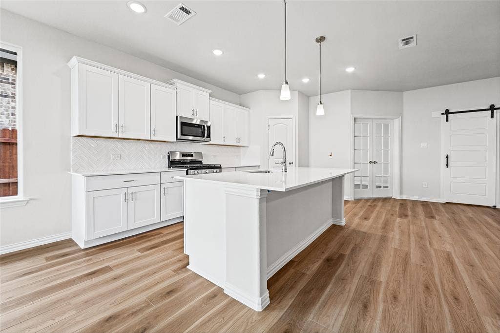 10004 Birchpoint Drive, Fort Worth, TX 76108 Modern kitchen featuring white cabinetry, stainless steel appliances, a center island with pendant lighting, wood-finish flooring, and a sliding barn door