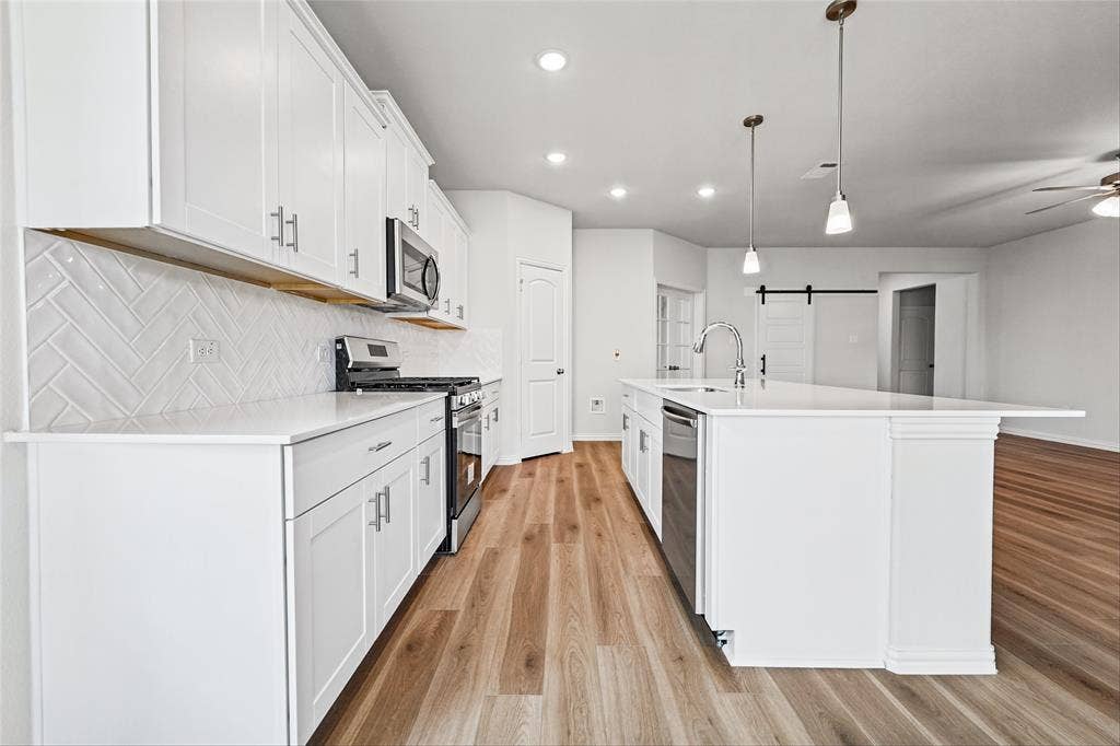 10004 Birchpoint Drive, Fort Worth, TX 76108 Kitchen featuring white shaker cabinetry, stainless steel appliances, a white tile herringbone backsplash, a large center island with a sink, and wood-finish flooring