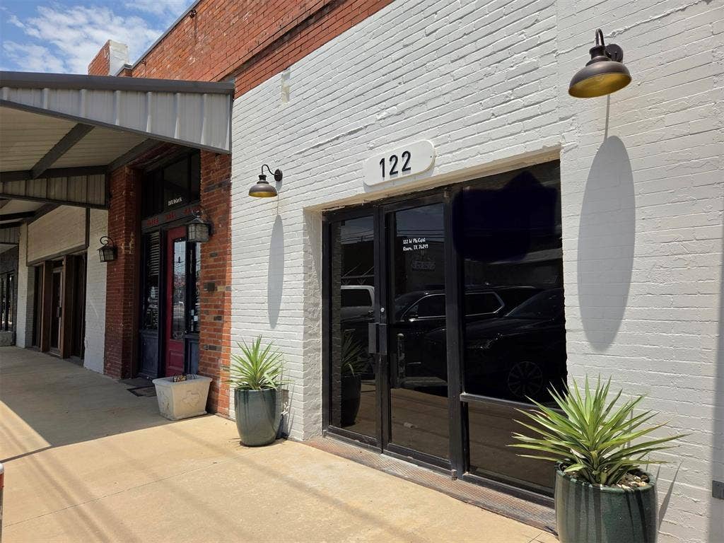 122 W Mccart Street, Krum, TX 76249 Commercial storefront featuring a brick facade with a white painted finish, large glass double doors, and two exterior wall-mounted light fixtures