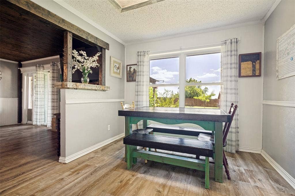 906 Clearview Street, Krum, TX 76249 Dining area featuring LVP flooring, a large window, half-wall paneling, crown molding, and a pass-through opening with a wood beam accent