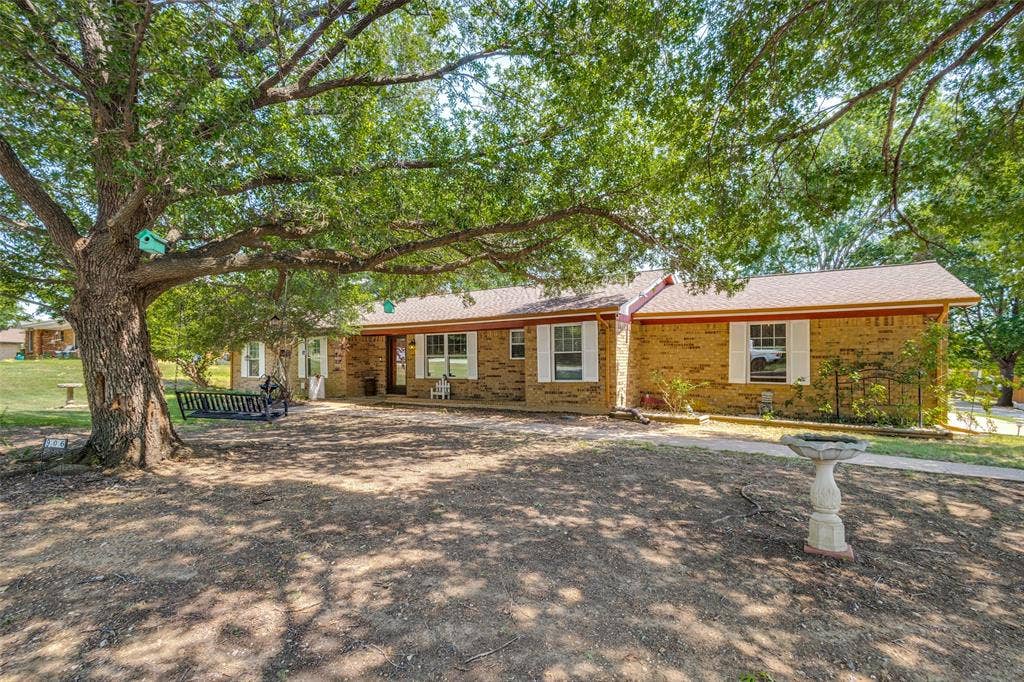 906 Clearview Street, Krum, TX 76249 Brick ranch-style residence featuring a large mature tree, a front porch, white window shutters.
