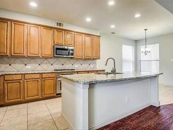 13990 Stevens Point Drive, Frisco, TX 75033 Kitchen featuring a central island with an integrated sink, granite countertops, wood cabinetry, stainless steel appliances, and tile backsplash