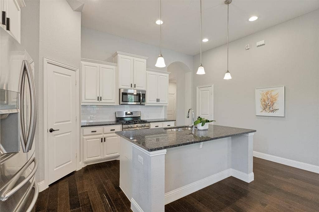 Kitchen featuring a central island with a sink, stainless steel appliances, white cabinetry, dark stone countertops, and wood-finish flooring