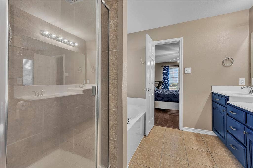1201 Destiny Court, Wylie, TX 75098 Primary bathroom featuring a spacious walk-in shower with glass enclosure and tile surround, dual vanities with blue cabinetry, ceramic tile flooring, and abundant lighting above the mirror
