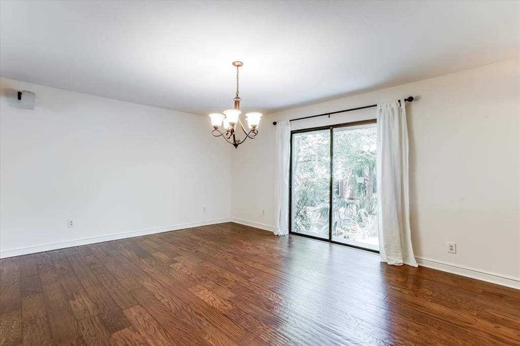 Spacious room featuring wood-finish flooring, a contemporary chandelier, white baseboards, and a large sliding glass door with white curtains