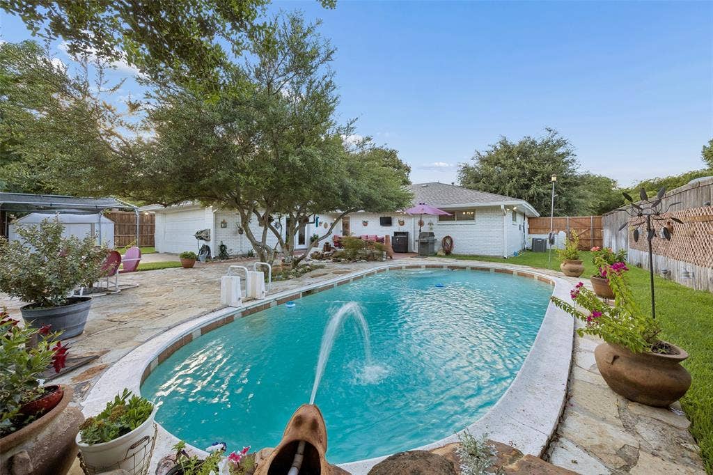 2104 Westlake Drive, Plano, TX 75075 Backyard oasis featuring an oval swimming pool with water features, extensive stone patio, mature trees, and a white brick exterior