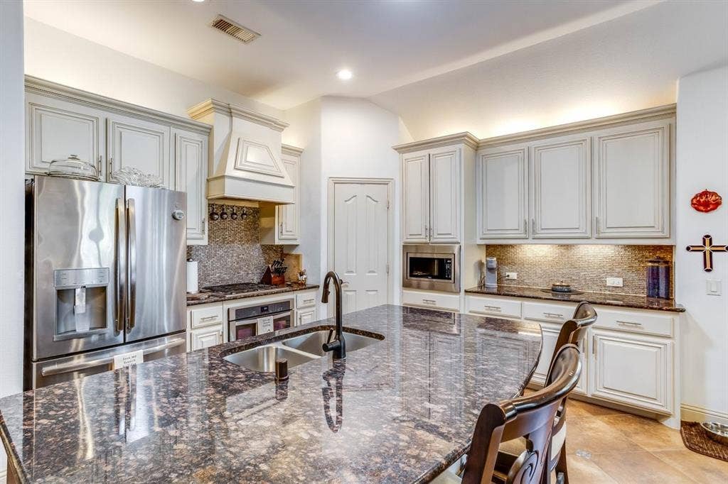 114 Lyndhurst Drive, Wylie, TX 75098 Kitchen featuring an expansive island with a dual-basin sink, under-cabinet lighting, stainless steel appliances, a decorative range hood, and light-toned cabinetry