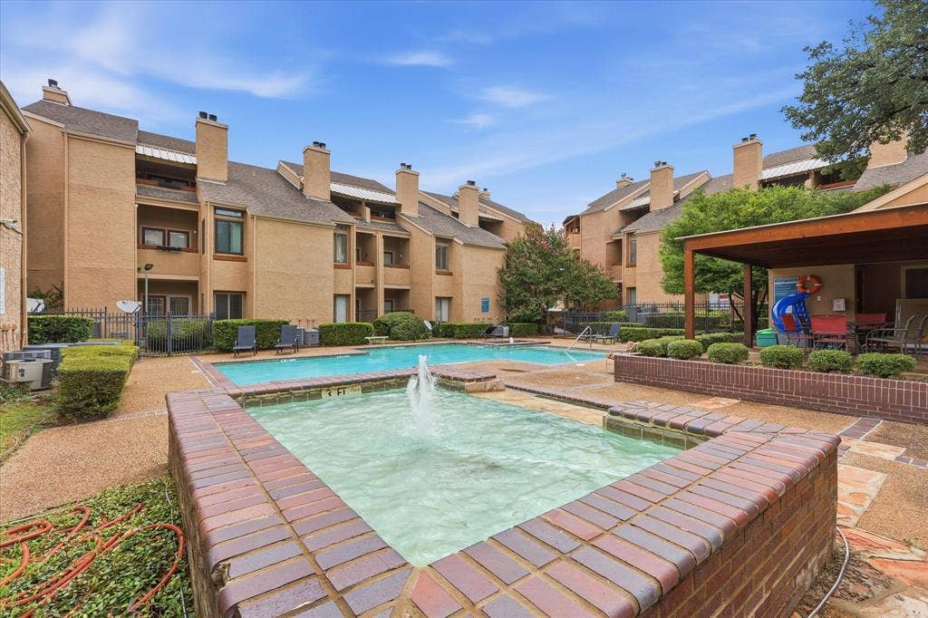 5550 Spring Valley Road, #A31, Dallas, TX 75254 Community pool area featuring a brick-lined fountain, a swimming pool with a 3-foot depth marker, a covered patio structure, and surrounding mature landscaping