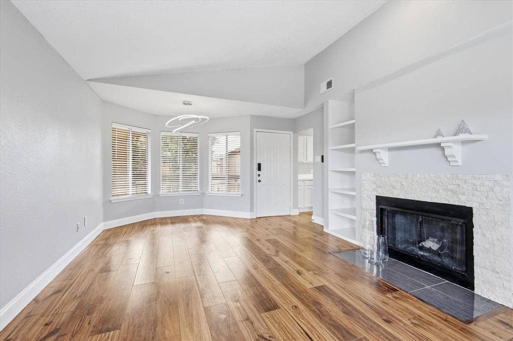 5550 Spring Valley Road, #A31, Dallas, TX 75254 Spacious living area featuring wood-finish flooring, a fireplace with a stone surround, built-in shelving, a modern ceiling light fixture, and multiple windows with blinds