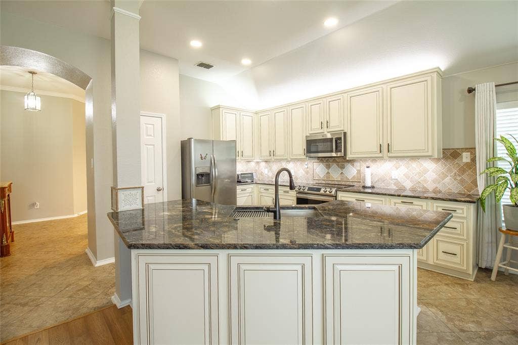 406 Hackberry Drive, Fate, TX 75087 Kitchen featuring an island with a black countertop and an integrated sink, cream-colored cabinetry, stainless steel appliances, and mosaic tile backsplash