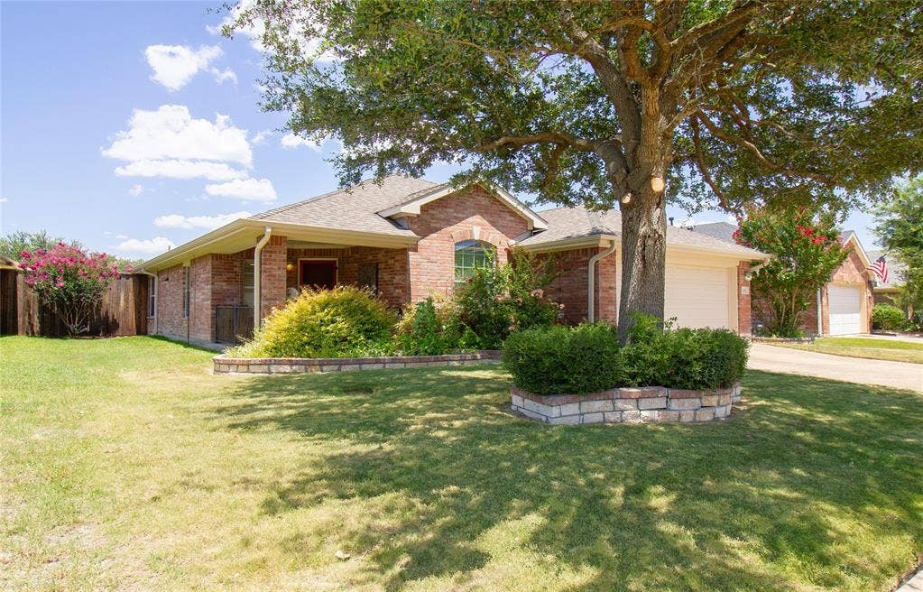 406 Hackberry Drive, Fate, TX 75087 Brick exterior residence featuring a prominent mature tree, landscaped beds with stone block edging, a covered front entry, and an attached garage with a white door