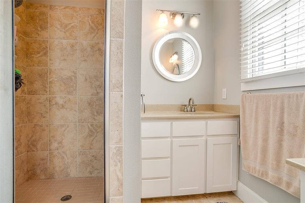 1211 S Adams Street, Fort Worth, TX 76104 Bathroom featuring a tiled standing shower, white vanity with a light-toned countertop, round wall-mounted mirror, and a window with blinds