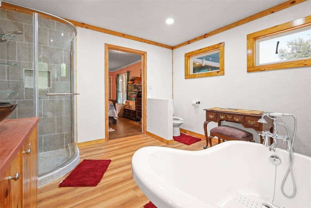 28300 County Road 2100, Kemp, TX 75143 Bathroom featuring a freestanding soaking tub, wood-finish flooring, a curved glass shower enclosure with gray tile, and wood-framed windows