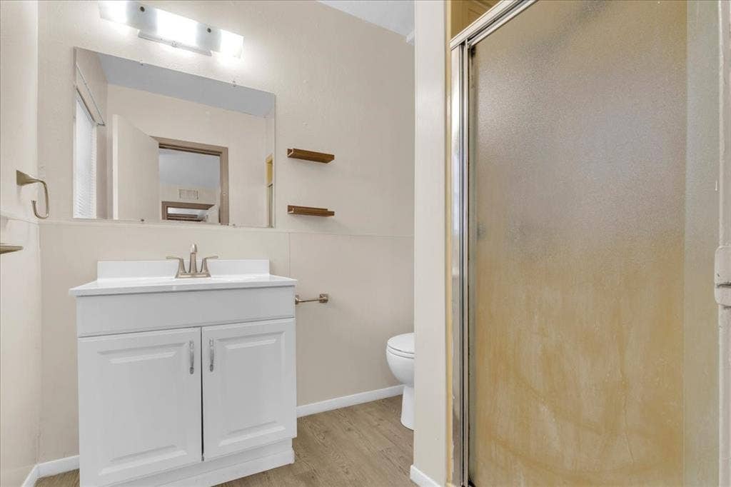 1113 Hudgins Avenue, Fort Worth, TX 76111 Bathroom featuring a single vanity with a white cabinet, a wall-mounted mirror, a two-bulb vanity light, and light wood-finish flooring