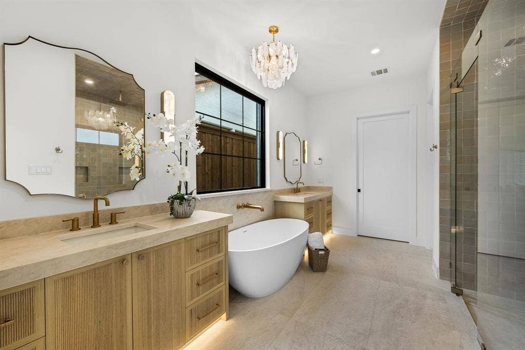 3723 Manana Drive, Dallas, TX 75220 Spacious bath featuring dual wood-finish vanities with light-toned countertops, a freestanding soaking tub, a large walk-in shower with glass enclosure, and a statement chandelier