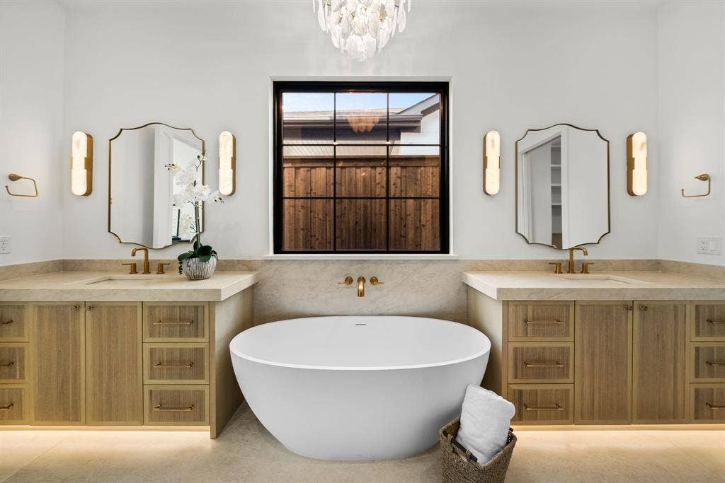 3723 Manana Drive, Dallas, TX 75220 Spa-inspired primary bathroom featuring dual wood-finish vanities with integrated storage, dual framed mirrors with integrated sconce lighting, a freestanding soaking tub, heated flooring, and a black-framed window with interior shutters overlooking the landscape