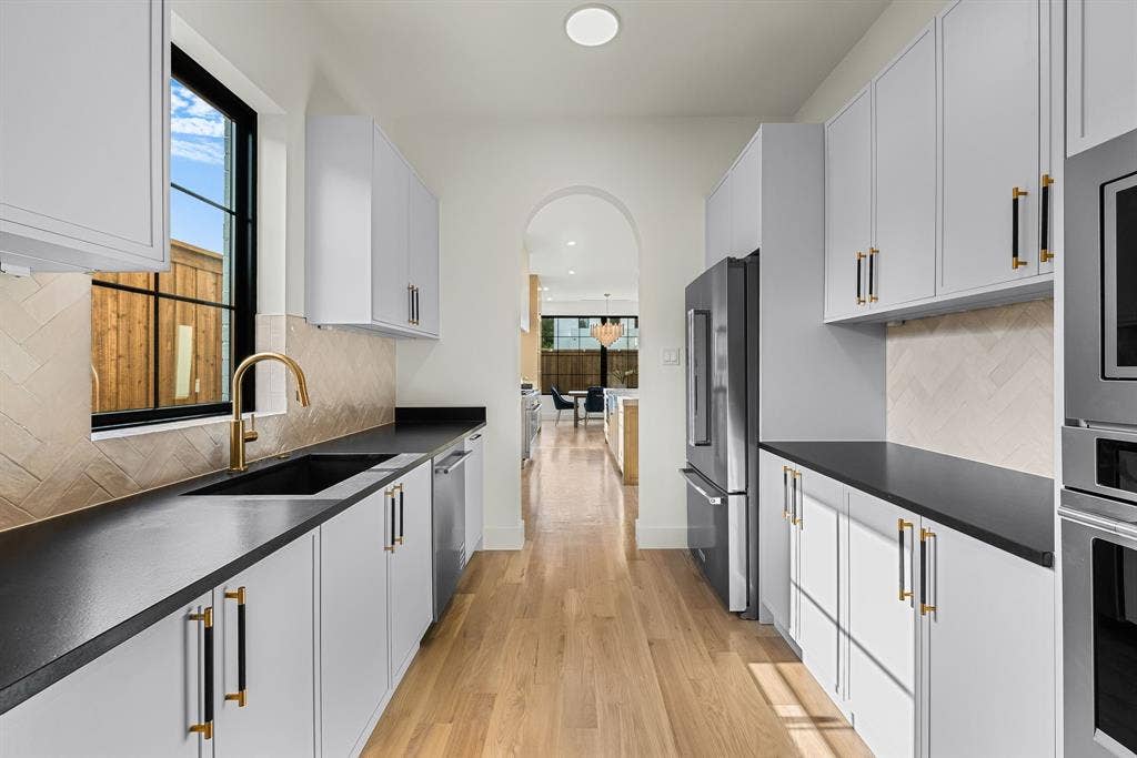 3723 Manana Drive, Dallas, TX 75220 Modern galley kitchen featuring light wood-finish flooring, white cabinetry with contrasting hardware, dark countertops, a herringbone tile backsplash, and a black-framed window