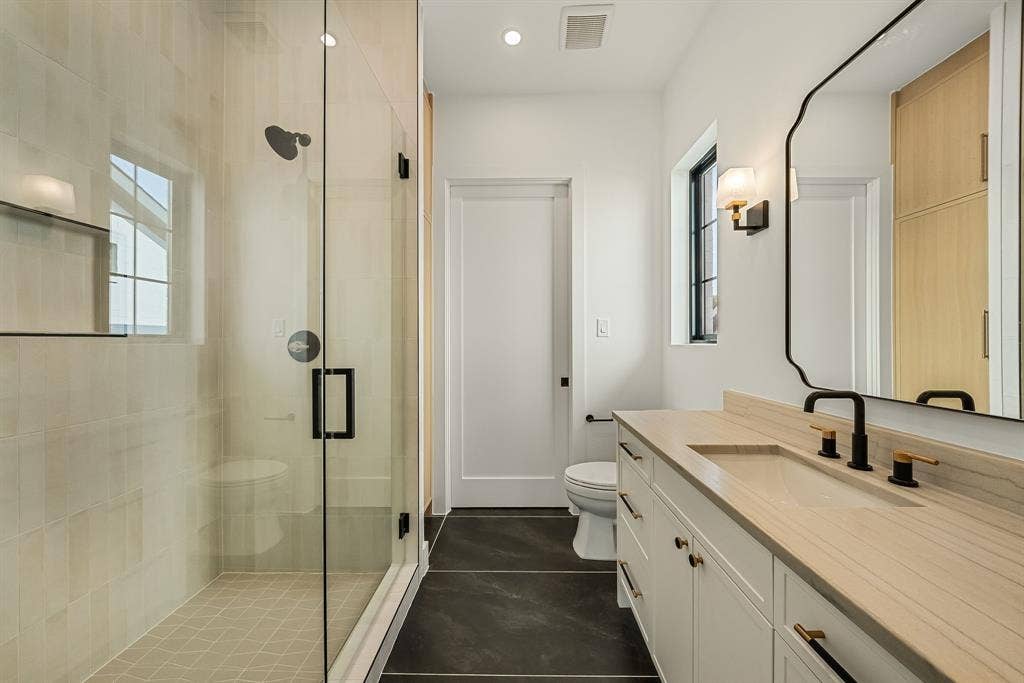 3723 Manana Drive, Dallas, TX 75220 Modern bathroom featuring a frameless glass shower enclosure, a vanity with an integrated sink and light wood-finish countertop, dark tile flooring, a black-framed window, and a decorative wall mirror