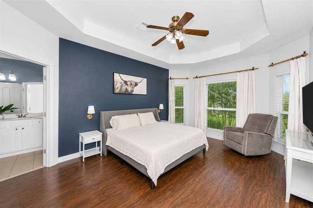 5304 Fox Run Drive, Watauga, TX 76137 Retreat to the spacious primary suite featuring a dramatic tray ceiling, rich engineered hardwood flooring, abundant natural light from multiple windows, and a striking accent wall. Notice the custom lighting on each side of the bed.
