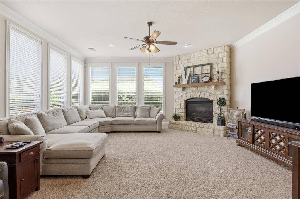 5030 Monroe Court, Midlothian, TX 76065 Living area featuring a stone fireplace with a wood mantel, light-colored carpeting, a ceiling fan, crown molding, and multiple windows with horizontal blinds