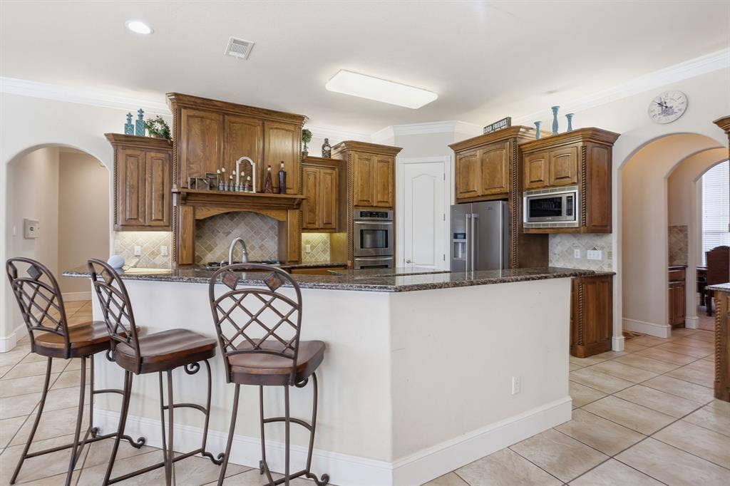5030 Monroe Court, Midlothian, TX 76065 Kitchen featuring wood cabinetry, stainless steel appliances, a tiled backsplash, and a granite-look island with a breakfast bar