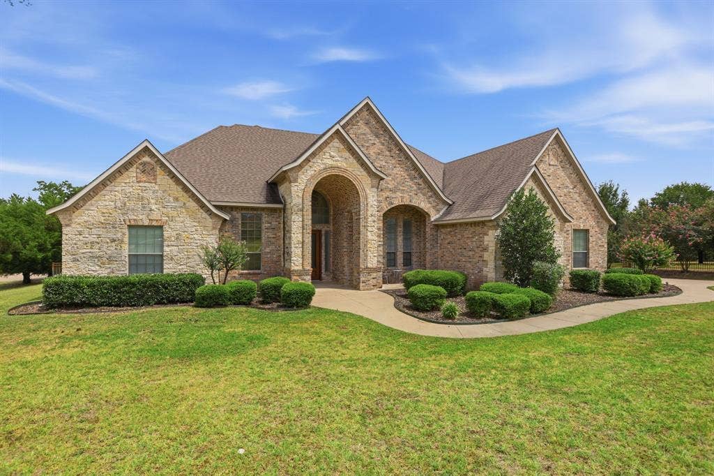 5030 Monroe Court, Midlothian, TX 76065 Stone facade residence featuring a prominent arched entry, a gabled roofline, a curved concrete walkway, and established foundation plantings