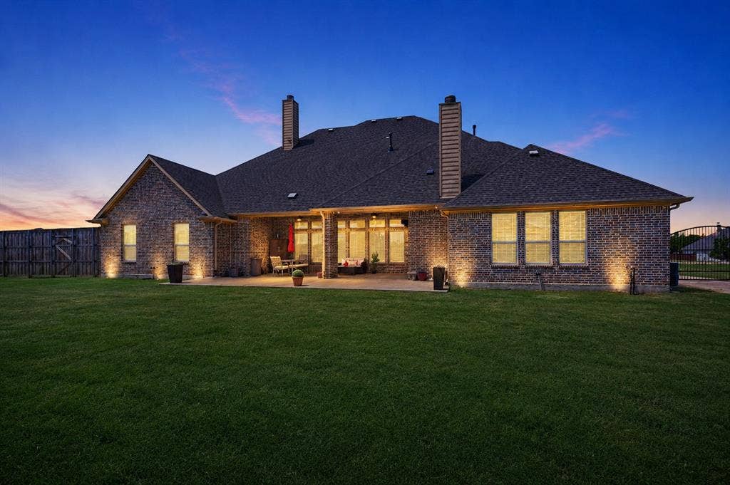 Expansive backyard with a manicured lawn, a full brick exterior, a covered patio, multiple windows, and exterior accent lighting