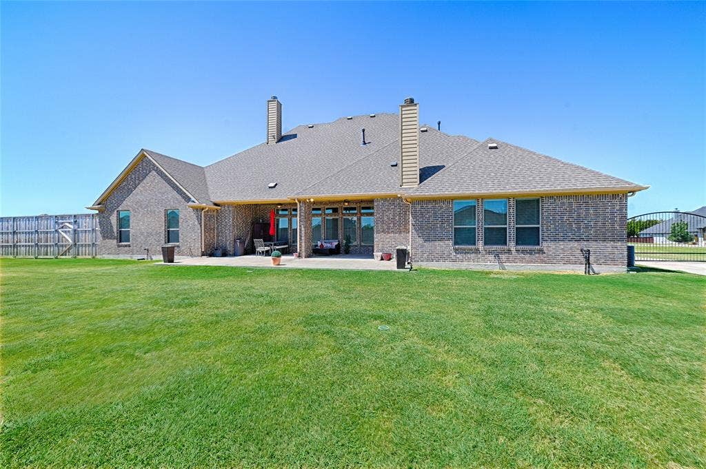 Spacious backyard featuring an extended patio, a large expanse of green lawn, brick exterior, multiple windows, and a shingle roof