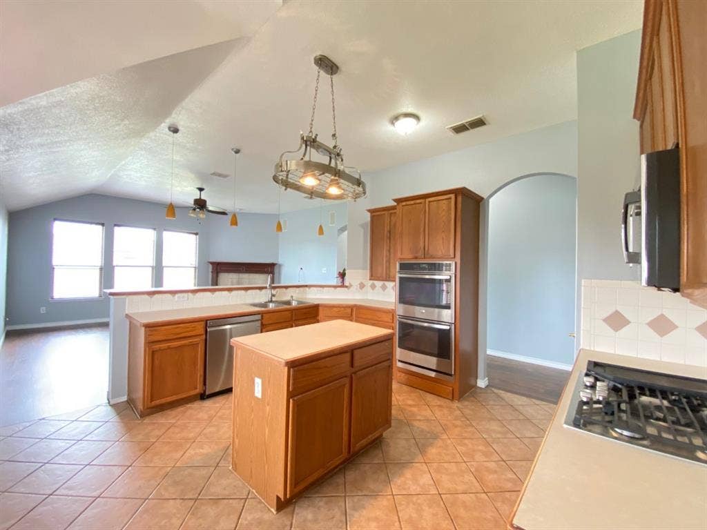 1328 Falcon Hollow, Cedar Hill, TX 75104 Spacious kitchen featuring tile flooring, wood cabinetry, a center island, stainless steel appliances including double ovens and a dishwasher, and an arched doorway leading to an additional room with vaulted ceilings and a fireplace