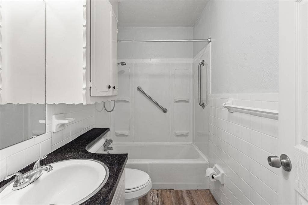 3407 Highmeadow Lane, Greenville, TX 75402 Full bathroom featuring a white tub/shower combination with grab bars, a vanity with a dark countertop and undermount sink, a toilet, and wood-finish flooring