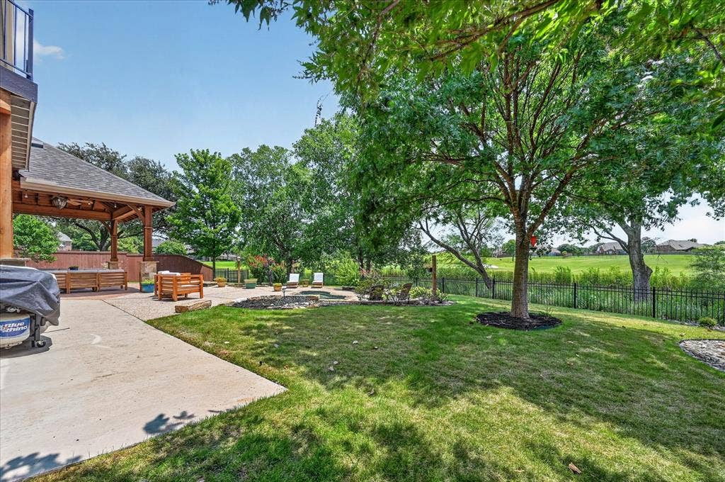 8405 Gallery Way, McKinney, TX 75072 Expansive backyard featuring a large concrete patio, a covered outdoor living area with wood beams, a stone-edged fire pit, mature trees, and a black wrought-iron fence
