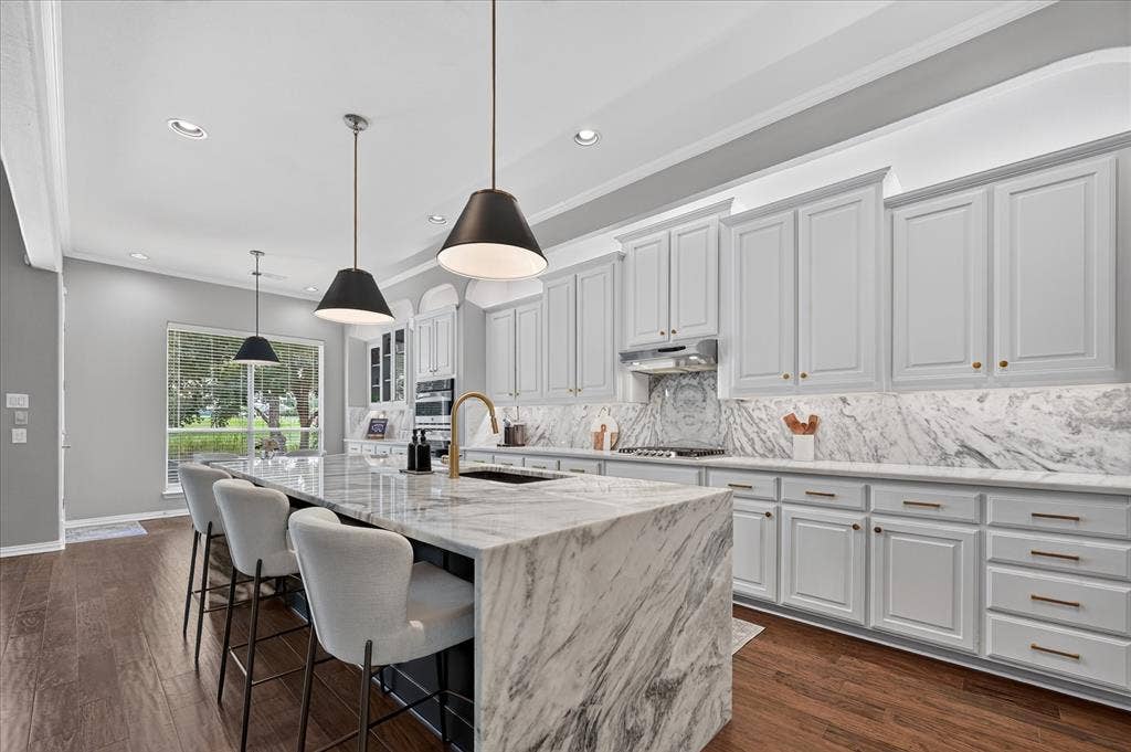 8405 Gallery Way, McKinney, TX 75072 Gourmet kitchen featuring a waterfall island with stone countertop, white cabinetry with gold-tone hardware, full-height stone backsplash, and dark wood-finish flooring
