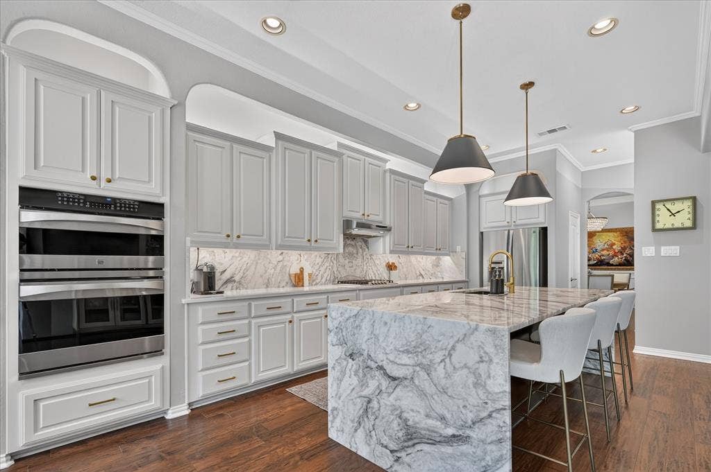 8405 Gallery Way, McKinney, TX 75072 Gourmet kitchen featuring a waterfall island with stone countertop, stainless steel double ovens, integrated cabinetry, recessed lighting, and wood-finish flooring