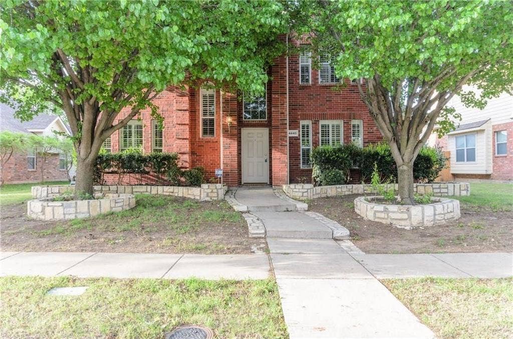 4440 Waskom Drive, Plano, TX 75024 Brick facade residence featuring a central entryway with a white door, framed by two established trees with stone-lined planters