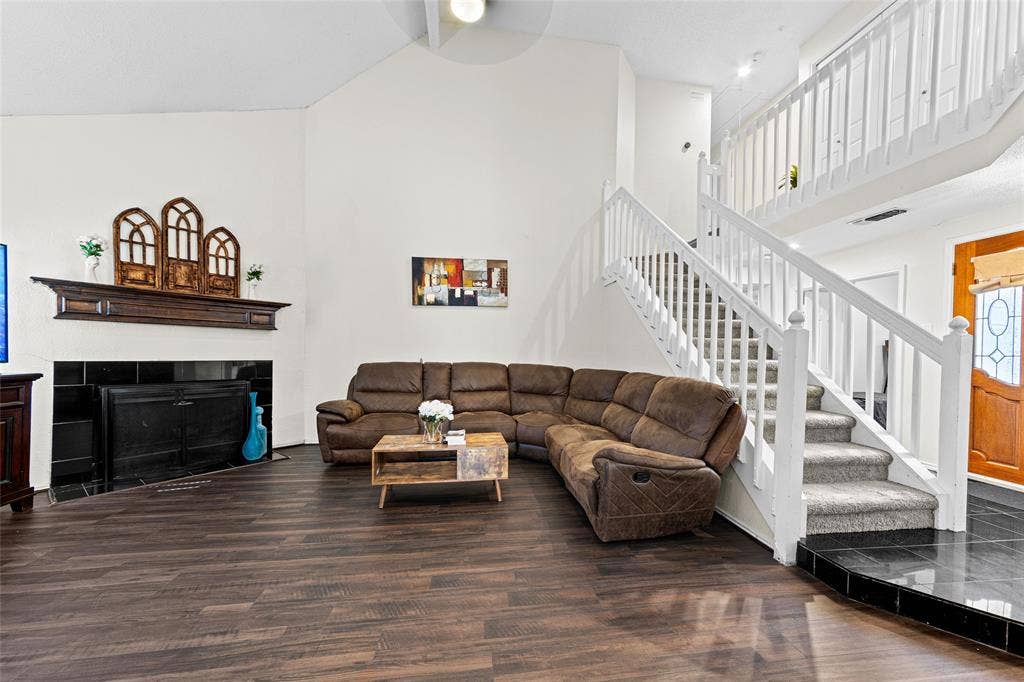 Spacious living area featuring wood-finish flooring, a white vaulted ceiling, and a fireplace with a dark tile surround and a wood mantel