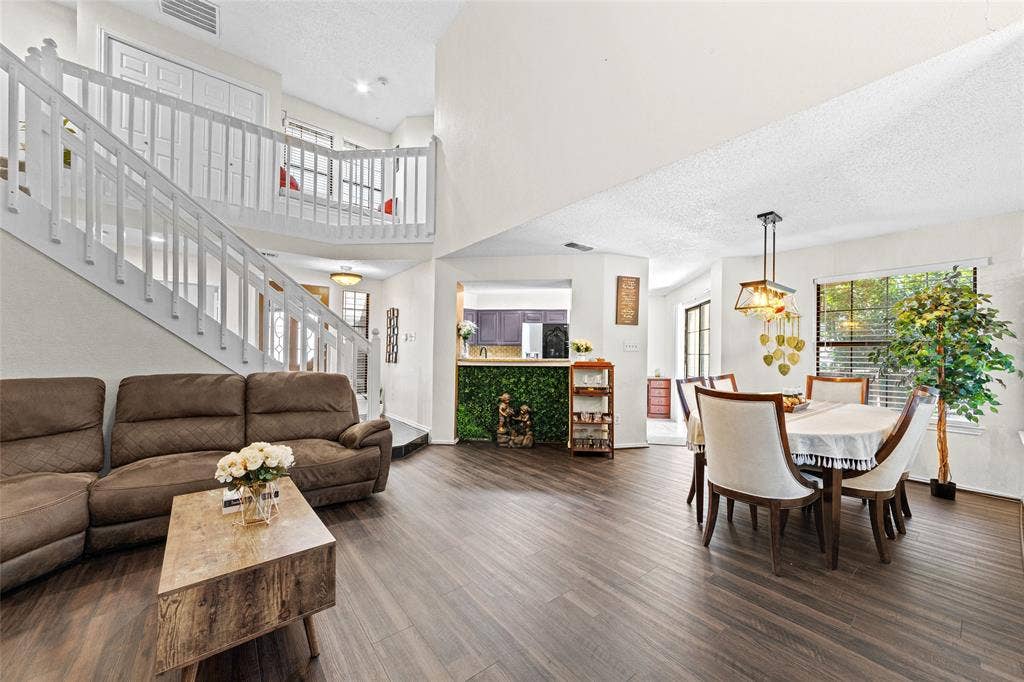 Open-concept living area featuring high ceilings, wood-finish flooring, a prominent staircase with white railings, and a dining area with an overhead light fixture
