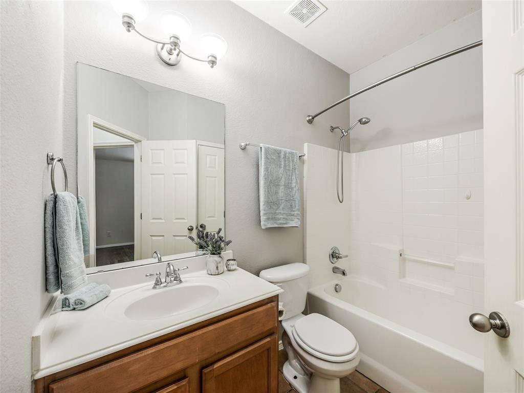 414 Dartmoor Drive, Celina, TX 75009 Full guest bathroom located at 414 Dartmoor Dr in Carter Ranch, Celina, TX. Includes a wood single-sink vanity with white countertop, 3-light vanity light fixture, large mirror, standard toilet, shower/tub combination with white tile surround, and neutral tile flooring.