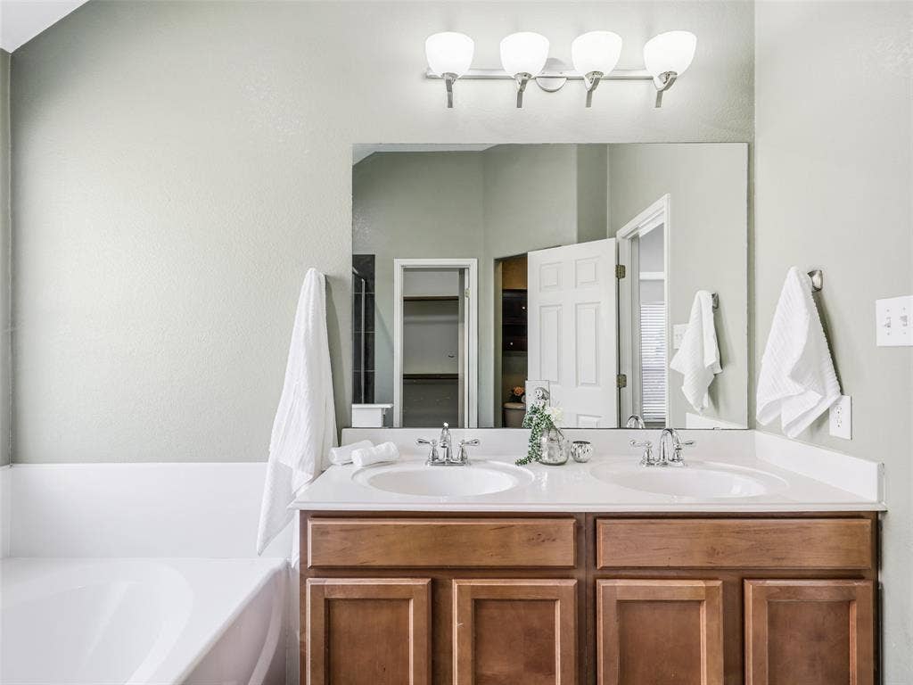 414 Dartmoor Drive, Celina, TX 75009 Primary ensuite bathroom at 414 Dartmoor Dr in Carter Ranch. Features a double vanity with wood cabinetry and dual undermount sinks, modern brushed nickel faucets, a wall-to-wall mirror, 4-light vanity fixture, a deep soaking garden tub.