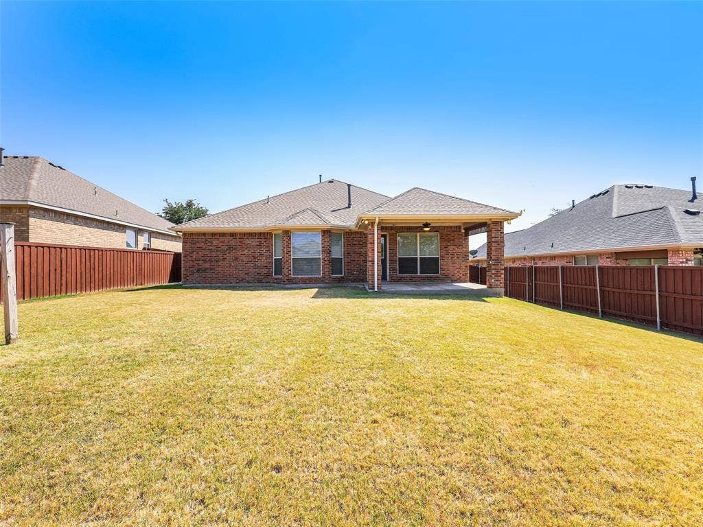 414 Dartmoor Drive, Celina, TX 75009 Centered shot from the back fence looking straight at the house at 414 Dartmoor Dr, Celina, TX. Highlights the architectural bay window pop-out from the primary suite, the covered patio structure, composite shingle rooflines, and a vast open lawn under clear blue Texas skies.