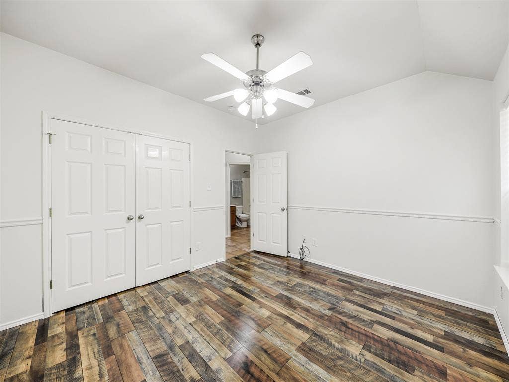 414 Dartmoor Drive, Celina, TX 75009 Reverse angle of the secondary bedroom at 414 Dartmoor Dr, Celina, Texas. Showcases the double-door closet, decorative chair rail trim, wood-look plank flooring, white ceiling fan, and direct doorway access to the adjacent guest hallway bathroom.