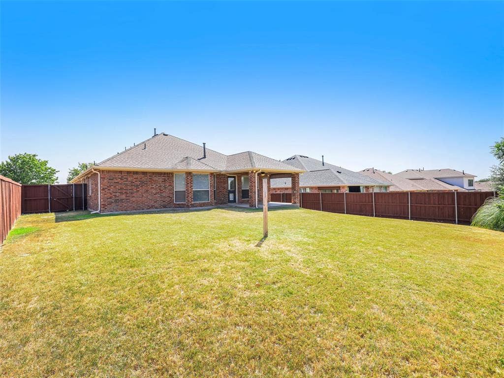 414 Dartmoor Drive, Celina, TX 75009 Straight-on perspective of the rear exterior at 414 Dartmoor Dr in Celina's Carter Ranch neighborhood. Showcases the single-story all-brick construction, covered patio extension with ceiling fan, side gate access, and fully enclosed wood privacy fence.