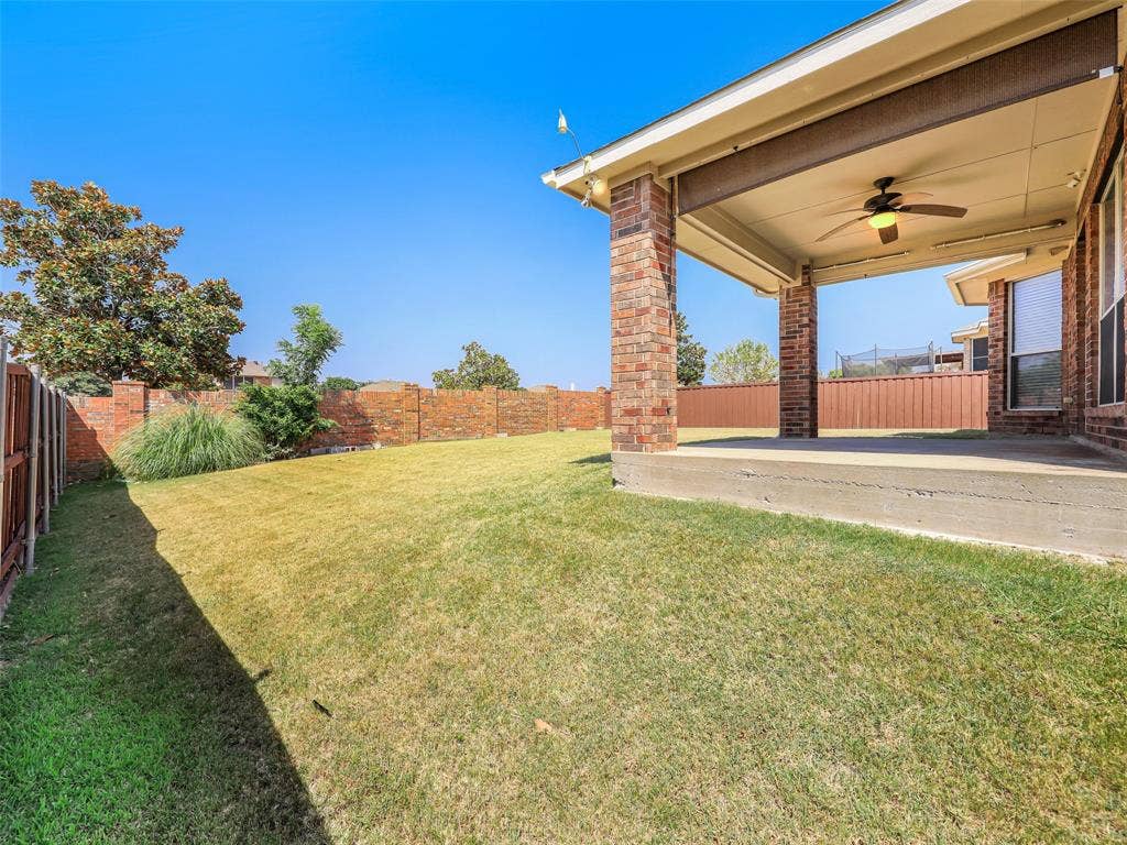 414 Dartmoor Drive, Celina, TX 75009 An exterior view of the covered back patio at 414 Dartmoor Dr, Celina, TX. Features classic brick support pillars, an outdoor ceiling fan with light fixture, a concrete slab foundation, a pull-down outdoor privacy shade, and a view looking out onto the spacious grassy backyard enclosed by wooden and brick perimeter fencing.