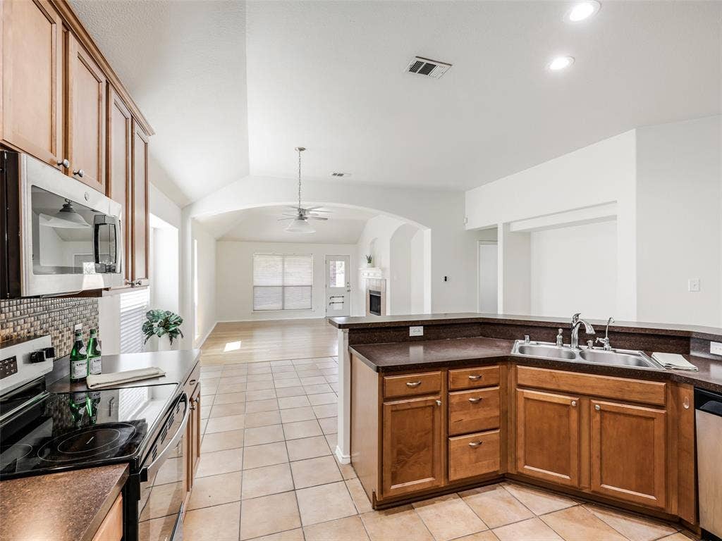 414 Dartmoor Drive, Celina, TX 75009 A interior view from inside the kitchen at 414 Dartmoor Dr in Celina's Carter Ranch neighborhood. The kitchen features warm wood cabinetry, a raised breakfast bar, a stainless steel double sink with a pull-down faucet, and neutral ceramic tile flooring. The space opens seamlessly into the main living room, which features a corner fireplace and vaulted ceilings.