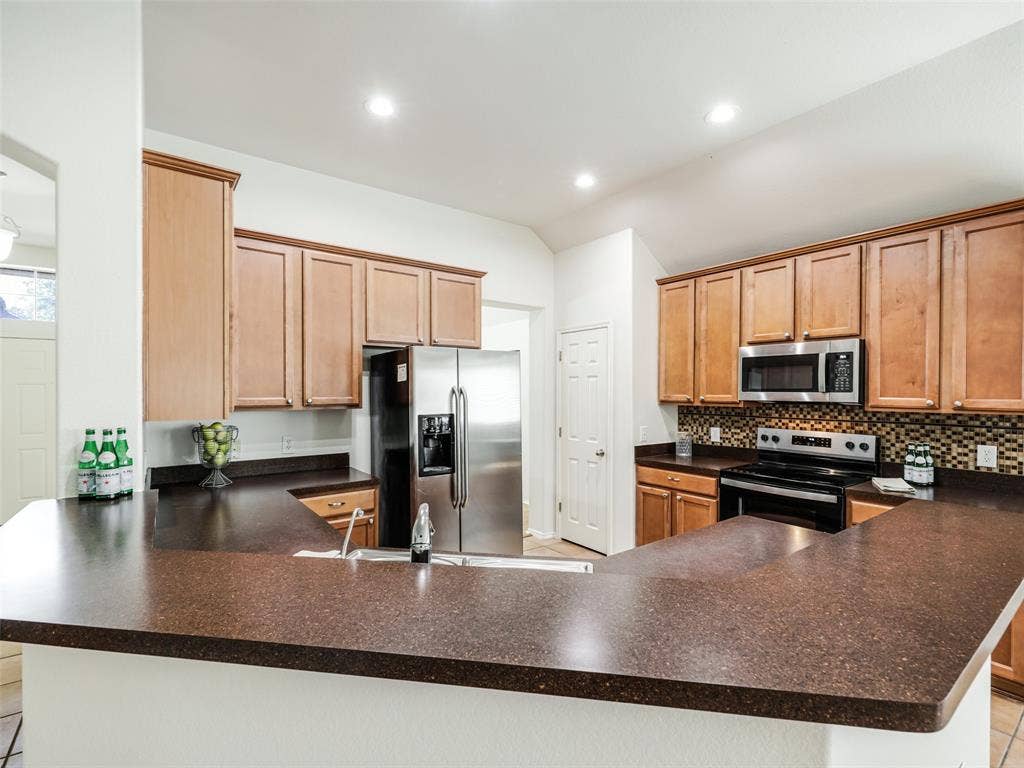 414 Dartmoor Drive, Celina, TX 75009 An open-angle view showcasing the wrap-around breakfast bar and kitchen footprint at 414 Dartmoor Dr in Carter Ranch. Highlighting upper and lower wood cabinets, dark stone-look countertops, glass mosaic backsplash tile, recessed lighting, and tile flooring, this layout provides ample counter space and seating capacity for entertaining.