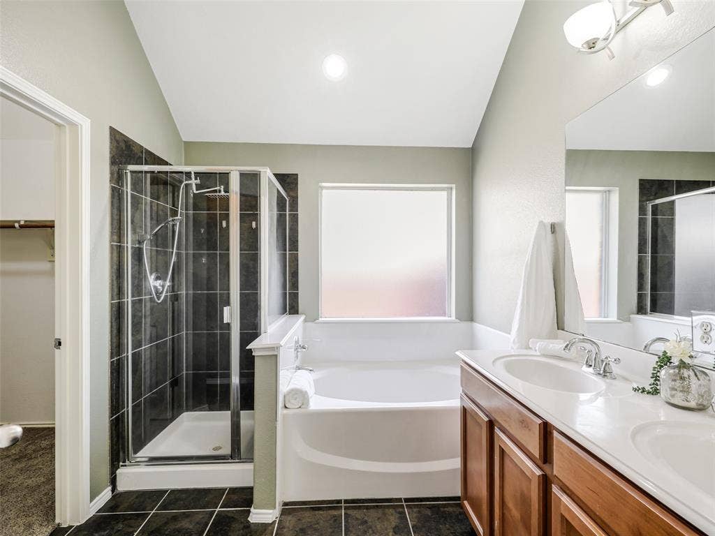 414 Dartmoor Drive, Celina, TX 75009 Bathroom featuring a glass-enclosed shower with dark tile surround, a soaking tub with frosted windows, a double vanity with wood cabinetry and dual sinks, and dark tile flooring